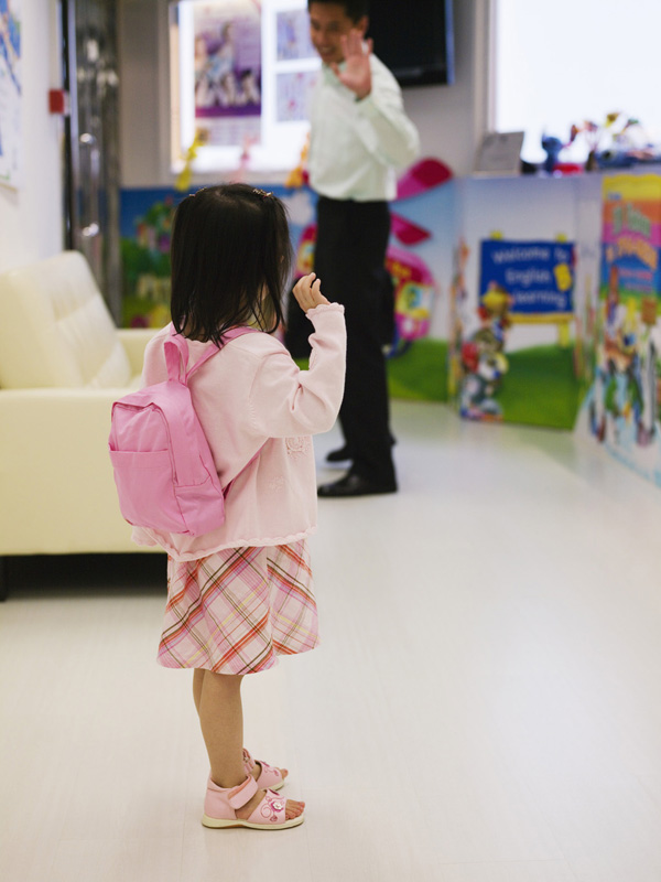 Little girl wearing a backpack waves goodbye to her father as he drops her off at preschool.